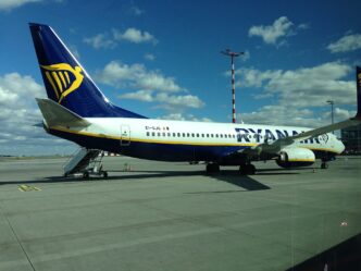 Boeing 737 v tmavě modrých barvách Ryanairu stojí na stojánce u nástupních schůdků, v pozadí obloha s mraky.