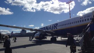 Cestující jdou po odbavovací ploše k tmavě modrému Boeingu 737 s bílým nápisem Ryanair na trupu.