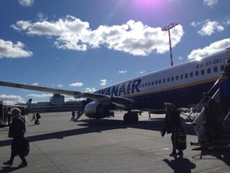Cestující jdou po odbavovací ploše k tmavě modrému Boeingu 737 s bílým nápisem Ryanair na trupu.
