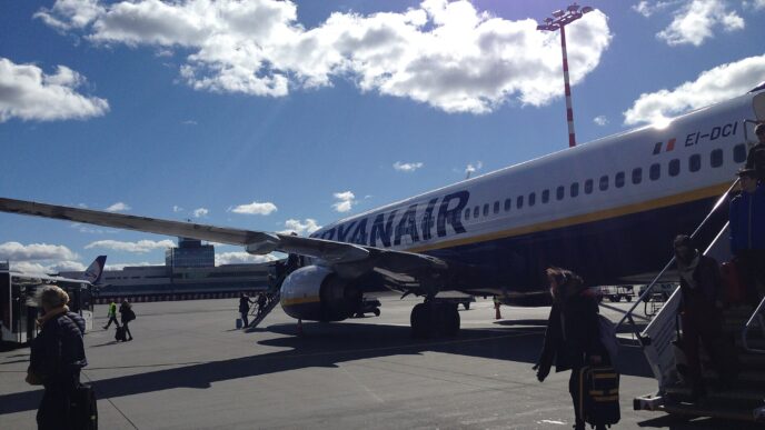 Cestující jdou po odbavovací ploše k tmavě modrému Boeingu 737 s bílým nápisem Ryanair na trupu.