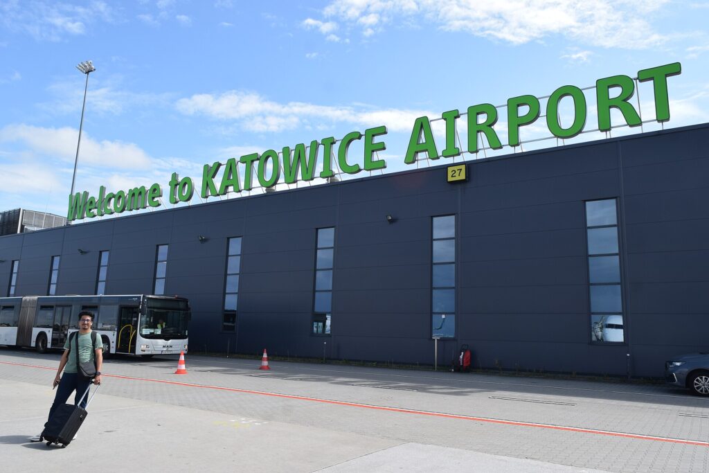 Fasáda odletového terminálu letiště Katovice-Pyrzowice s nápisem Welcome to Katowice Airport, před ní autobus a cestující s kufrem