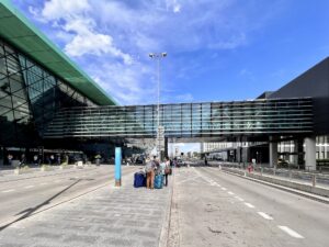 Prosklená fasáda terminálu Kraków Airport s krytou lávkou nad příjezdovou komunikací a cestujícími s kufry před vchodem