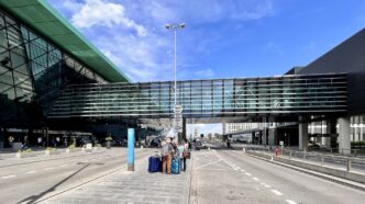 Prosklená fasáda terminálu Kraków Airport s krytou lávkou nad příjezdovou komunikací a cestujícími s kufry před vchodem