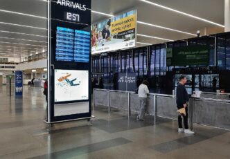 Příletová hala Terminálu 1 pražského letiště s informační tabulí odletů a cestujícími u přepážek