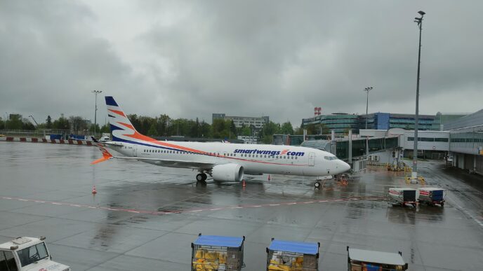 Boeing 737 MAX 8 v modro-oranžových barvách Smartwings stojí na mokré odbavovací ploše, v popředí zavazadlové vozíky.